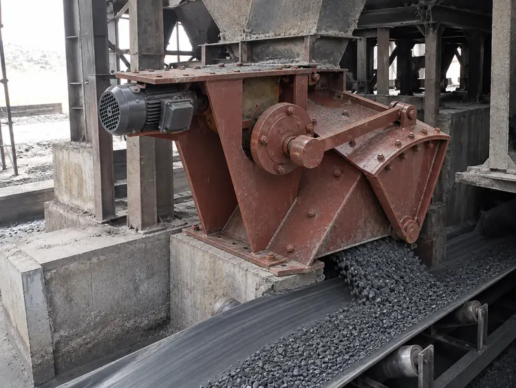 Red-brown feeder beneath a storage hopper discharging dark material onto a belt conveyor
