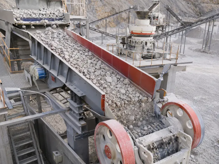 Gray vibrating feeder with red inner sidewalls delivering rocks toward a crusher