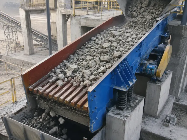 Blue grizzly feeder carrying gray rocks beneath a hopper inside an industrial plant